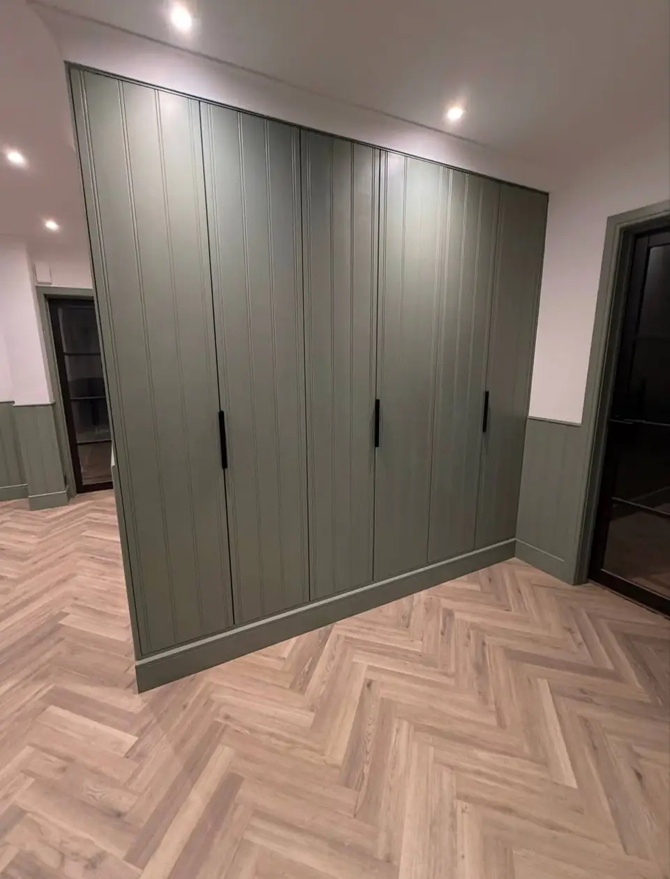 Full-height green fitted cupboards above panelled walls on a herringbone floor