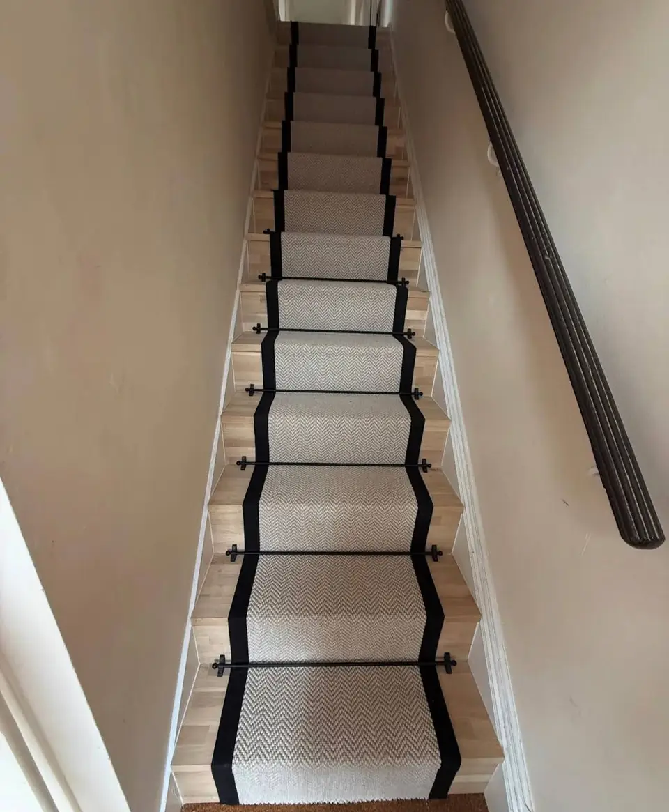 Narrow staircase with a cream runner, black bound edges and black stair rods, dark handrail on the wall