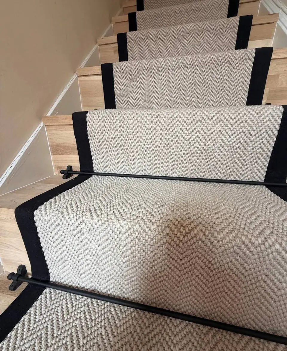 Close-up of a herringbone stair runner with black bound edges held by a black stair rod